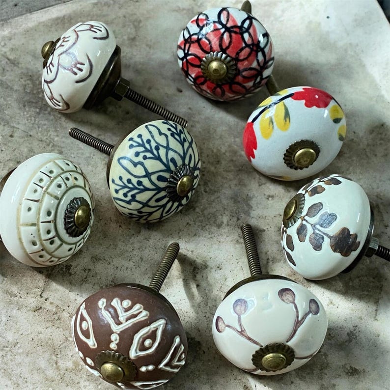Puede incluir: Una colecci&oacute;n de ocho pomos de cer&aacute;mica para armarios con varios dise&ntilde;os pintados a mano. Los pomos presentan una mezcla de colores, incluyendo blanco, marr&oacute;n, rojo y azul, con herrajes de lat&oacute;n. Cada pomo tiene un patr&oacute;n &uacute;nico, como floral, geom&eacute;trico y abstracto.