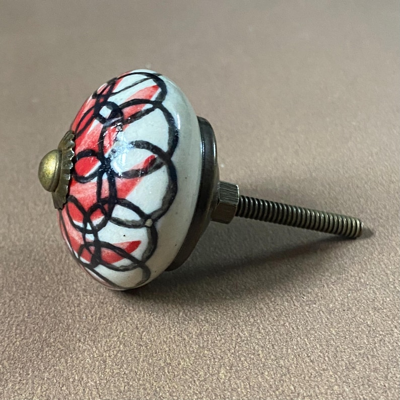 Puede incluir: Pomo de armario de cer&aacute;mica con base blanca, adornado con un dise&ntilde;o abstracto rojo y negro. Tiene un acento dorado y un tornillo met&aacute;lico para la fijaci&oacute;n. Ideal para a&ntilde;adir un toque distintivo a los muebles, con un di&aacute;metro de unos 4 cm.