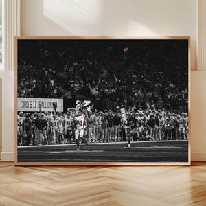 May include: Black and white framed print of a football game in action. The image captures a moment of play with players in uniforms, a crowd in the background, and the text "3RD & 11 BALL ON" visible. The print is displayed on a wooden floor.