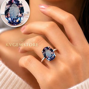 Peut inclure: Une bague en argent avec une grande pierre précieuse ovale à changement de couleur, passant de nuances de bleu profond à violet. La monture comporte quatre griffes maintenant la pierre. Le texte KVGEMSTORE est visible sur l'image.