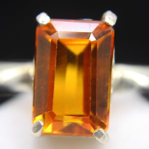 Könnte beinhalten: Ein Silberring mit einem großen, rechteckig geschliffenen, bernsteinfarbenen Edelstein. Der Edelstein hat eine brillante, facettierte Oberfläche, die das Licht reflektiert. Das Ringband ist schlicht und aus poliertem Silber.