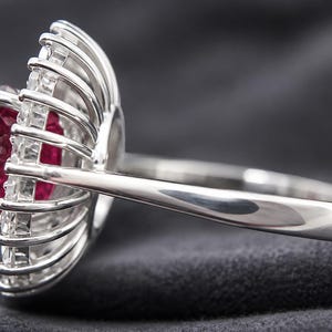 May include: A silver-toned ring featuring a central rectangular red gemstone surrounded by a halo of small, clear, baguette-cut stones arranged in a radial, sunburst-like pattern, set on a smooth, polished metal band with a side profile view.