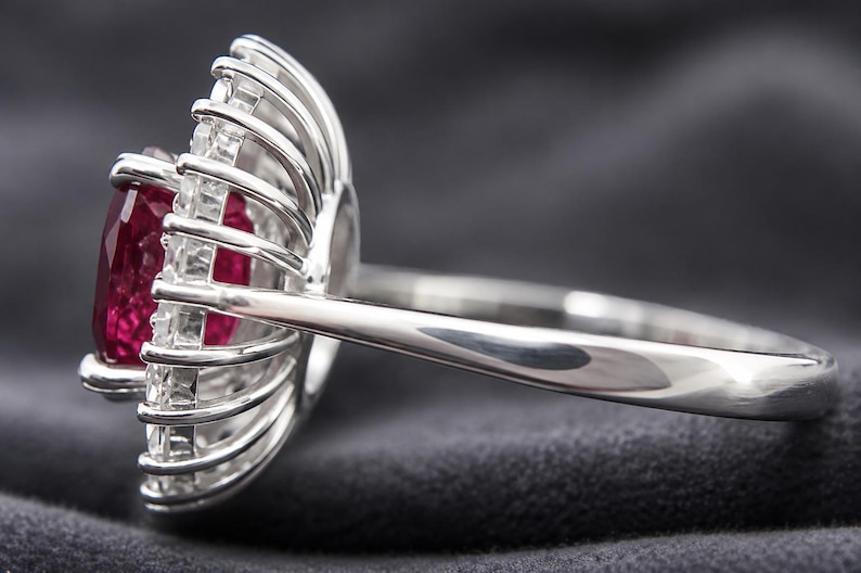 May include: A silver-toned ring featuring a central rectangular red gemstone surrounded by a halo of small, clear, baguette-cut stones arranged in a radial, sunburst-like pattern, set on a smooth, polished metal band with a side profile view.