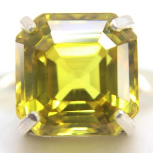 May include: A close-up of a square-cut, yellow-green gemstone set in a silver ring. The gemstone has multiple facets that reflect light, creating a brilliant sparkle. The ring's setting features four prongs that secure the stone.