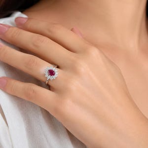 May include: A silver-toned ring featuring a central pear-shaped deep red gemstone surrounded by a halo of small, clear, round-cut stones. The ring has a slender, polished band and is worn on a finger, showcasing the intricate floral-like setting.