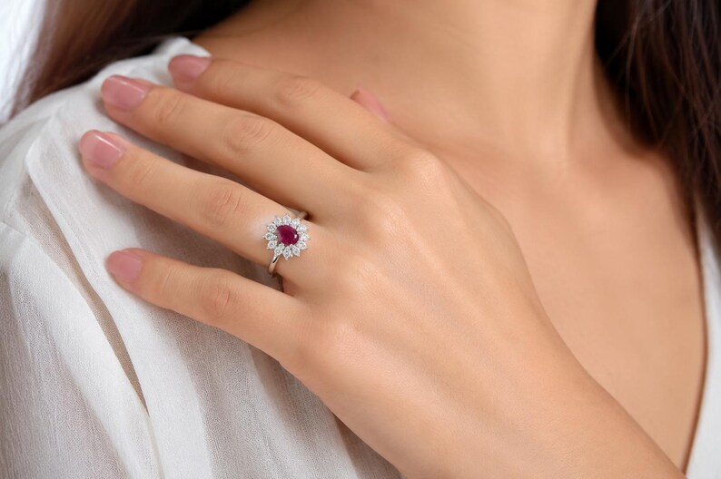 May include: A silver-toned ring featuring a central pear-shaped deep red gemstone surrounded by a halo of small, clear, round-cut stones. The ring has a slender, polished band and is worn on a finger, showcasing the intricate floral-like setting.