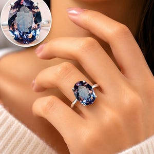 Op de afbeelding: Een zilveren ring met een grote, ovaal geslepen edelsteen die van kleur verandert tussen diepblauw, paars en roodachtig roze tinten. De steen is gezet in een zetting met vier pootjes op een eenvoudige, gepolijste zilveren band, getoond in een inzet.