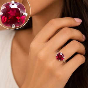 May include: A silver ring featuring a large, cushion-cut, deep red gemstone held in place by four prongs. The stone has a faceted surface that reflects light, and the ring is shown both on a finger and in a separate, enlarged circular inset view.