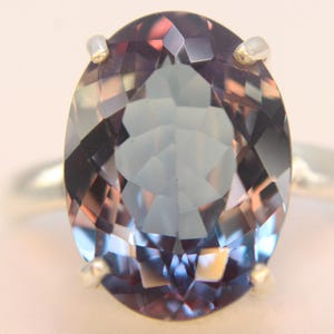 Può includere: Anello in argento con una gemma taglio ovale. La gemma presenta sfaccettature con sfumature di viola, blu e rosa. Le griffe dell'anello sono argentate. Sfondo morbido e neutro.