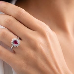 May include: A silver-toned ring featuring a central oval-cut deep red ruby gemstone surrounded by a halo of twelve small, round-cut clear white diamonds. The ring has a slender, polished metal band that sits securely on the finger.
