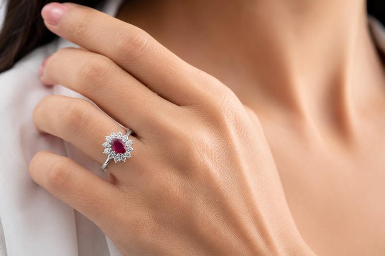 May include: A silver-toned ring featuring a central oval-cut deep red ruby gemstone surrounded by a halo of twelve small, round-cut clear white diamonds. The ring has a slender, polished metal band that sits securely on the finger.