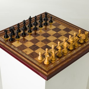 Könnte beinhalten: Ein Schachspiel aus Holz mit schwarzen und cremefarbenen Figuren, die auf einem karierten Brett angeordnet sind. Das Brett ist von einem dunklen Holzrahmen umgeben und steht auf einem weißen Sockel. Die Schachfiguren befinden sich in ihren Ausgangspositionen.