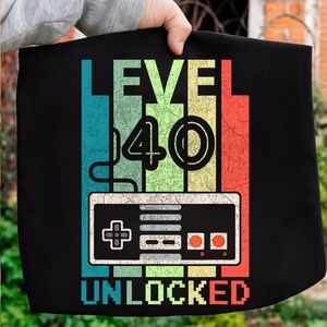 May include: Black graphic tee with the text "LEVEL 40 UNLOCKED". The design features a retro video game controller and colorful vertical stripes in teal, yellow, green, and red. The number 40 is prominently displayed.