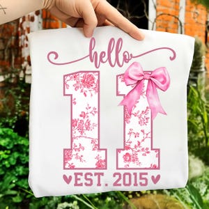 May include: White tote bag with the word "hello" in pink script above two pink floral-filled number 1s. A pink bow is on the right number 1. The text "EST. 2015" is below the numbers, with pink hearts on either side.