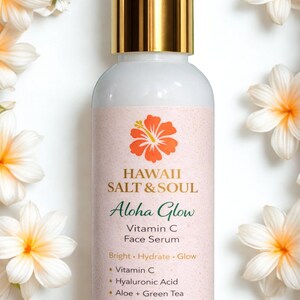 May include: A white bottle of Hawaii Salt & Soul Aloha Glow Vitamin C Face Serum with a gold cap. The label features a hibiscus flower and the text "Aloha Glow". The bottle is surrounded by white and yellow flowers.