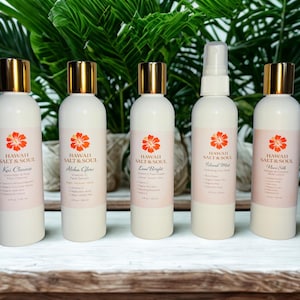 May include: Five white bottles of Hawaii Salt & Soul skincare products with gold caps and orange flower logos. The bottles are labeled: Kou Cleanse, Aloha Glow, Lave Bright, Island Mist, and Flower Silk. The background features green foliage.