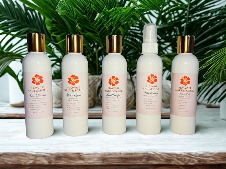 May include: Five white bottles of Hawaii Salt & Soul skincare products with gold caps and orange flower logos. The bottles are labeled: Kou Cleanse, Aloha Glow, Lave Bright, Island Mist, and Flower Silk. The background features green foliage.