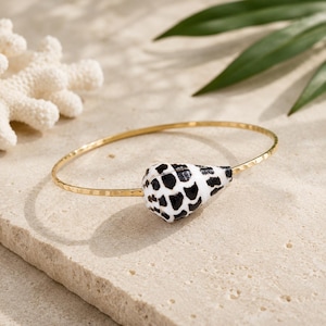 May include: A gold-toned metal bangle bracelet featuring a hammered texture and a single cone-shaped seashell as a focal point. The seashell is white with a distinct pattern of irregular black spots covering its entire surface.