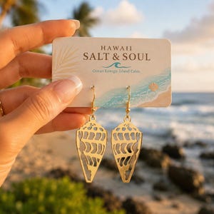 Puede incluir: Un par de pendientes colgantes de tono dorado con un diseño de concha recortada, suspendidos de una tarjeta de presentación con el texto HAWAII SALT & SOUL Ocean Energy. Island Calm. Los pendientes tienen una forma triangular y puntiaguda.