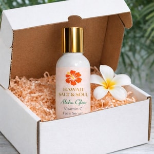 May include: A white gift box with a bottle of face serum, a white flower, and peach-colored packing material. The serum bottle has a gold cap and a label with the text "Hawaii Salt & Soul Aloha Glow Vitamin C Face Serum."