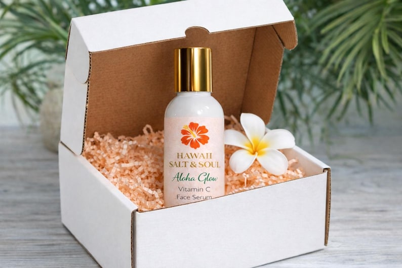 May include: A white gift box with a bottle of face serum, a white flower, and peach-colored packing material. The serum bottle has a gold cap and a label with the text "Hawaii Salt & Soul Aloha Glow Vitamin C Face Serum."