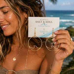 以下が含まれることがあります： 波の切り抜きデザインが施されたゴールドトーンのフープピアス。HAWAII SALT & SOUL, Ocean Energy, Island Calm, INSPIRED BY HAWAII MADE WITH ALOHAと印刷された白いディスプレイカードに掛けられています。
