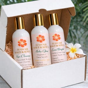 May include: A white gift box contains three white bottles with gold caps. Each bottle has a label with the words "Hawaii Salt & Soul" and a floral design. The bottles are labeled "Kai Cleanse", "Aloha Glow", and "Lani Bright".