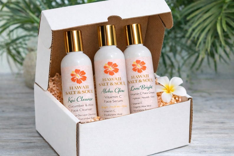 May include: A white gift box contains three white bottles with gold caps. Each bottle has a label with the words "Hawaii Salt & Soul" and a floral design. The bottles are labeled "Kai Cleanse", "Aloha Glow", and "Lani Bright".