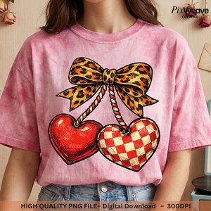May include: Pink t-shirt featuring a graphic of two heart-shaped cherries. One heart is solid red, the other is a red and white checkerboard pattern. A leopard print bow sits above the cherries. The shirt has the text "Wear" on the solid red heart.