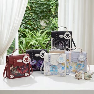 Puede incluir: Una colección de bolsos tipo satchel en varios colores, incluyendo burdeos, negro, blanco y azul claro. Cada bolso presenta un panel frontal transparente que muestra ilustraciones y un charm en forma de pata. Tienen asas superiores y correas ajustables.
