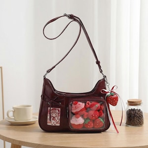 May include: A burgundy shoulder bag with a clear pocket filled with plush cupcakes and strawberries. A strawberry charm hangs from the strap. The bag has a small pocket with a cartoon image. A cup and saucer are on the table.