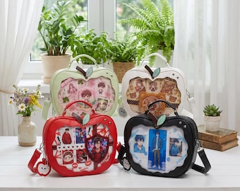 May include: Four apple-shaped handbags in pastel green, white, red, and black. Each bag has a clear window displaying character art and stickers. They feature a top handle, a detachable shoulder strap, and a leaf-shaped accent.