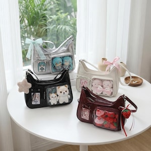 May include: Four shoulder bags in silver, black, cream, and burgundy, each with a clear pocket displaying plush toys. The bags have a bow or charm attached. The bags are on a white table near a window.