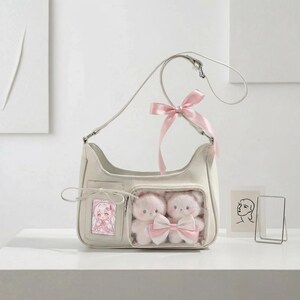 May include: Cream-colored shoulder bag with a clear pocket containing two plush teddy bears with a pink bow. A pink ribbon is tied to the strap. A small picture of an anime character is in a pocket on the front.
