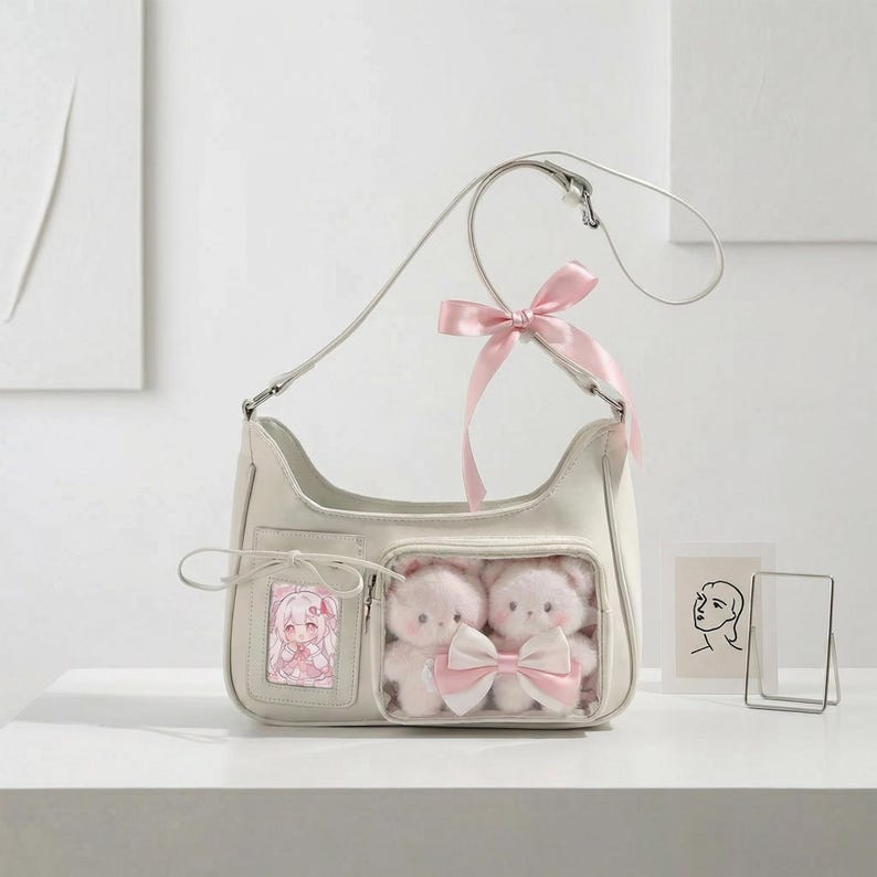 May include: Cream-colored shoulder bag with a clear pocket containing two plush teddy bears with a pink bow. A pink ribbon is tied to the strap. A small picture of an anime character is in a pocket on the front.