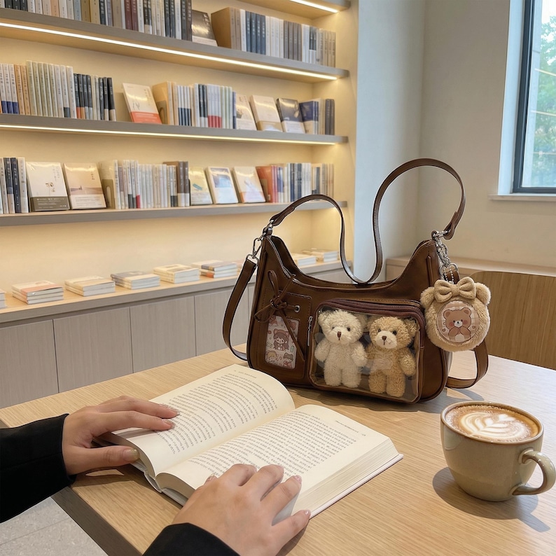May include: A brown shoulder bag with a clear pocket containing two teddy bears. The bag has a matching teddy bear keychain. A person is reading a book at a table with a coffee cup.