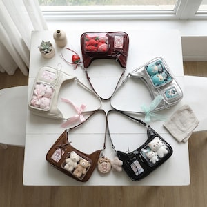 May include: An assortment of five shoulder bags in various colors, including white, brown, silver, and black. Each bag features a clear compartment filled with plush toys or decorative items. Some bags have decorative bows and charms.