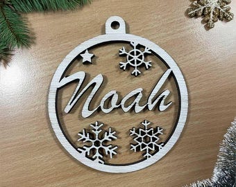May include: A wooden Christmas bauble with the name "Noah" cut out in a cursive font. The bauble is round with snowflake and star cutouts. The ornament is white with a dark border. Decorative greenery and a gold glitter snowflake are in the background.