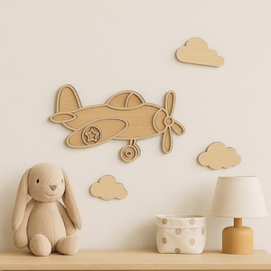 Puede incluir: Decoración de pared de avión de madera con tres recortes de nubes de madera. Un conejito de peluche y una pequeña lámpara con una pantalla beige se encuentran sobre una superficie de madera. El avión tiene una estrella en la rueda y una hélice.