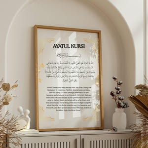 Puede incluir: Una impresión enmarcada con Ayatul Kursi en caligrafía árabe y traducción al inglés. La obra de arte tiene un borde dorado y se exhibe en un mueble blanco. Los elementos decorativos incluyen plantas secas y una pequeña escultura blanca.