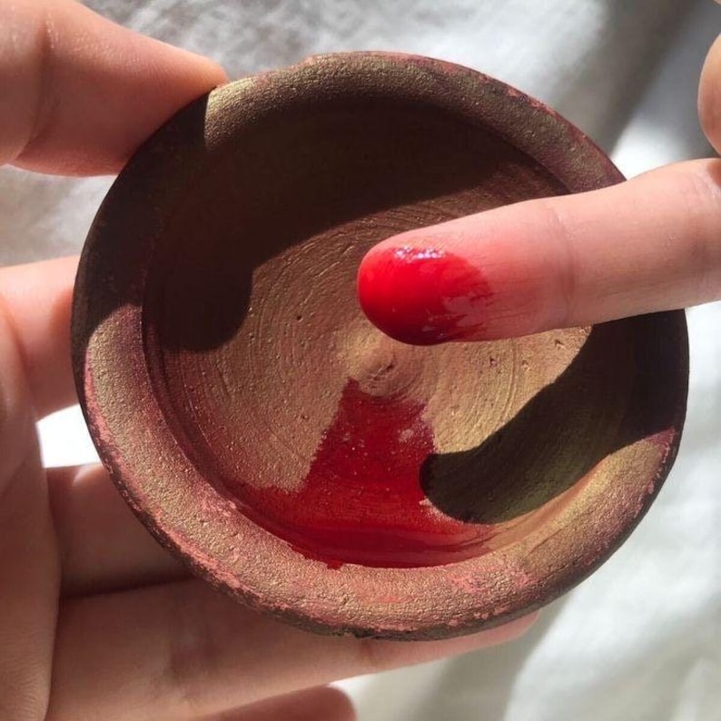 May include: A small, round, terracotta-colored bowl with a red liquid inside. A finger is dipped into the red liquid, showing the color and texture. The bowl has a gold-colored rim and interior.