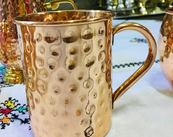 May include: A copper mug with a hammered texture and a curved handle. The mug has a shiny, reflective surface and is designed for beverages. The mug is part of a set, with other similar copper items visible in the background.