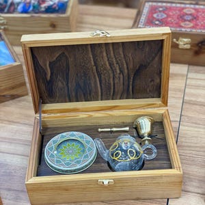 Może przedstawiać: Otwarta drewniana skrzynka z ciemnym drewnianym wnętrzem, zawierająca mały szklany czajniczek ze złotymi akcentami, okrągłą ozdobną puszkę, mały mosiężny kubek i mosiężną fajkę. Skrzynka jest zamknięta złotą klamrą.