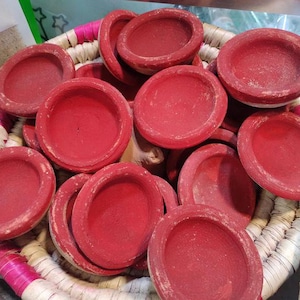May include: A collection of small, round, red clay bowls with a shallow design. The bowls are nestled in a woven basket with pink and white trim. The bowls have a matte finish and are likely handmade.