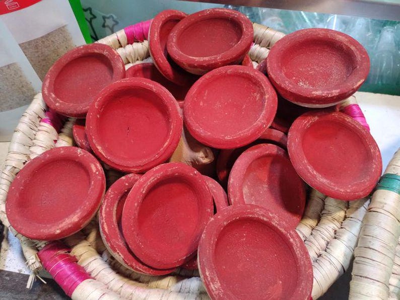 May include: A collection of small, round, red clay bowls with a shallow design. The bowls are nestled in a woven basket with pink and white trim. The bowls have a matte finish and are likely handmade.