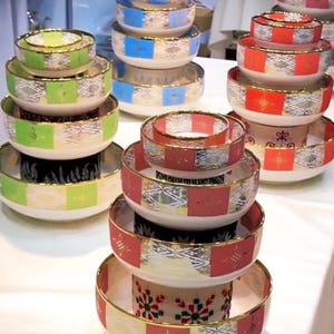 May include: A collection of decorative ceramic bowls in various sizes, stacked in sets. The bowls feature a white base with colorful accents of green, blue, and red squares, gold rims, and intricate patterns. The bowls are arranged on a white surface.