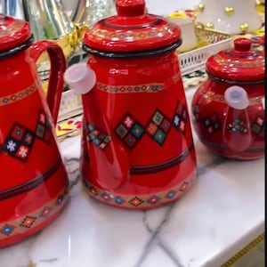 May include: Three red enamel teapots with matching lids and handles. The teapots have a traditional design with geometric patterns in blue, green, and white. The teapots are arranged on a white surface.