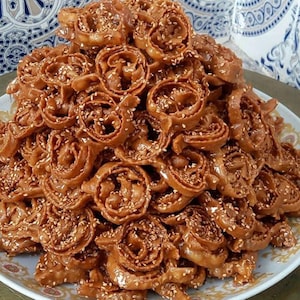 May include: A large pile of golden-brown, spiral-shaped pastries, generously sprinkled with sesame seeds, presented on a white plate with a decorative border. The pastries have a glossy finish, suggesting a sweet glaze.