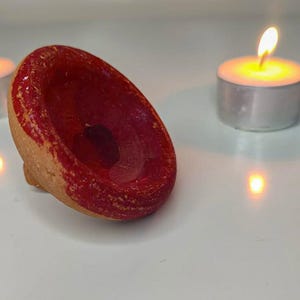 May include: A small, handmade ceramic bowl with a vibrant red interior and a natural, unglazed exterior. The bowl is set on a white surface, with a lit tea light candle in the background, creating a warm, inviting ambiance.
