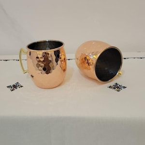 May include: Two copper Moscow Mule mugs with a hammered finish. One mug is upright, the other is on its side. Both have gold-coloured handles and dark interiors. The mugs are on a white tablecloth with embroidered details.